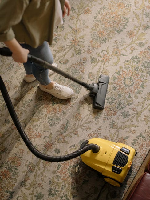 A person is vacuuming a patterned area rug with a beige background and floral motifs in shades of green, orange, and blue. The individual is wearing casual clothing including a tan jacket, dark jeans, and white sneakers, and is holding a vacuum cleaner with a long metal tube attached to a black vacuum head. The yellow and black canister vacuum cleaner is positioned on the carpet, with its hose connected to the head. The room appears well-lit, emphasizing the clean and dust-free condition of the rug, with the cleaning process focusing on surface dirt removal as part of domestic cleaning services provided by Cleaner Earls Court.