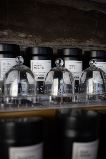 Close-up view of cylindrical black jars with white labels, arranged on a glass shelf in a retail setting, with clear glass domed lids placed upside down in front of them. The jars are labeled with dif