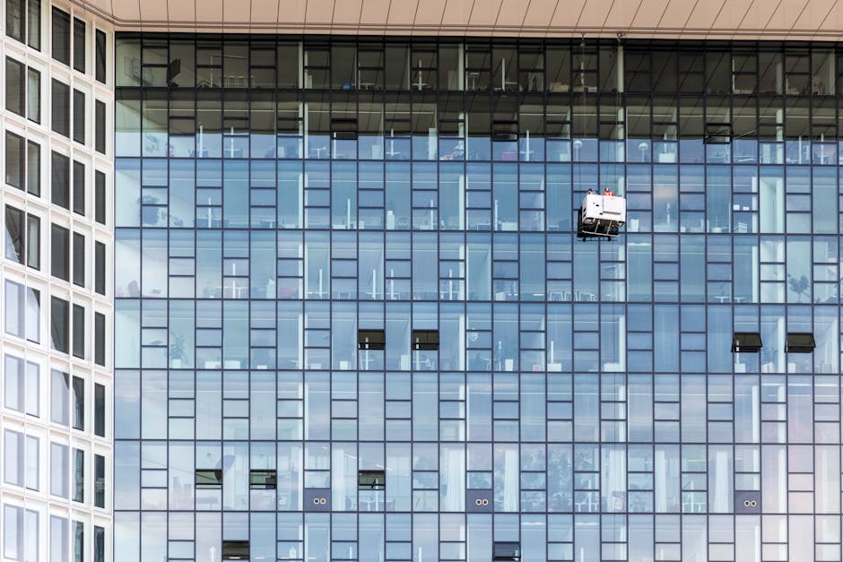 A close-up view of a modern high-rise building's exterior focusing on glass windows and a small balcony where two cleaning workers are performing surface cleaning and window maintenance. The building'